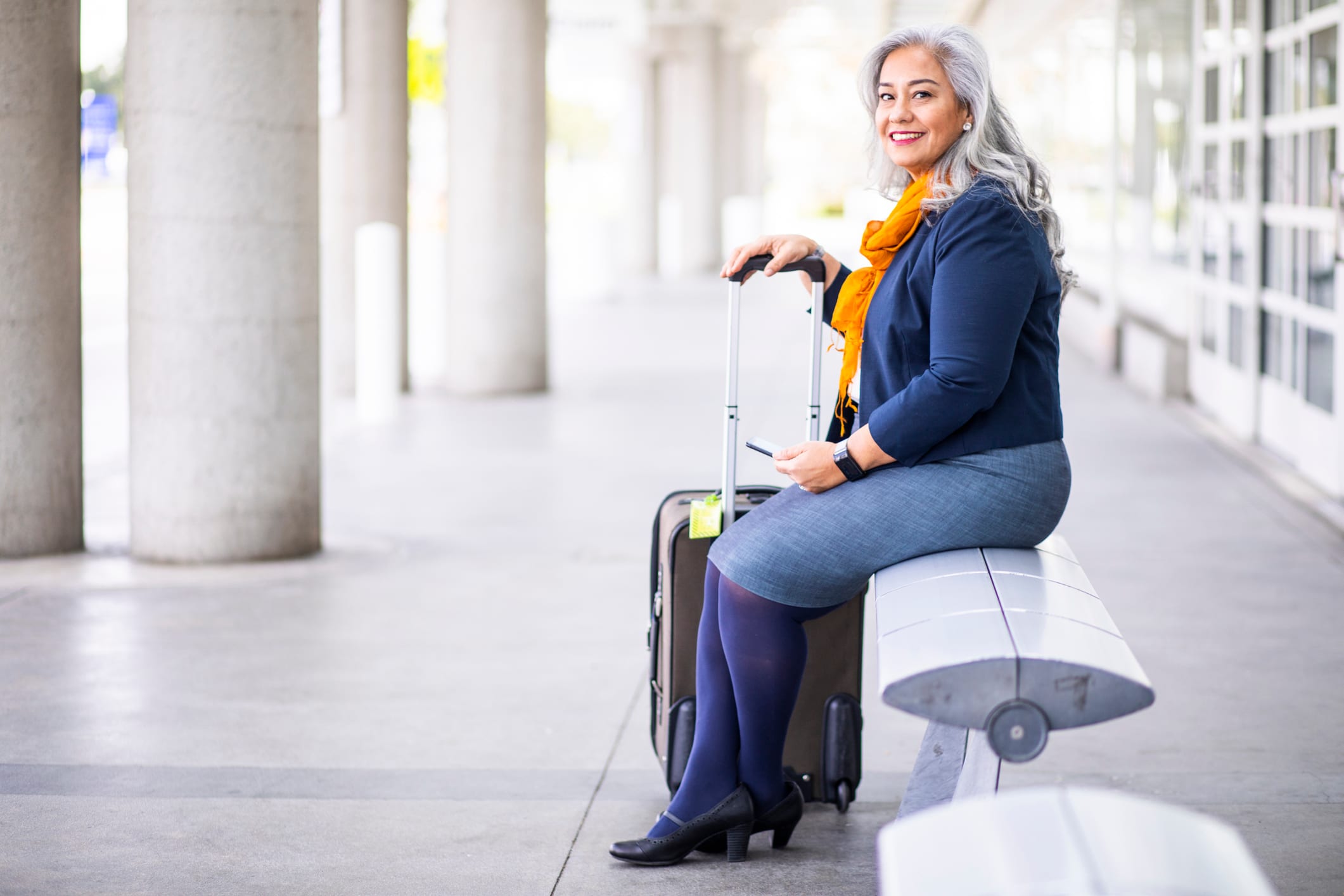 Femme aux cheveux gris assise à l’extérieur d’un aéroport.
